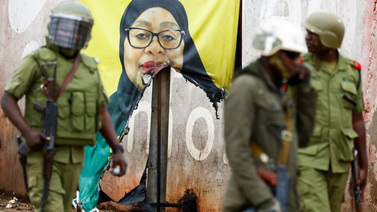 Tanzanian riot police officers walk past a vandalised campaign poster of President Samia Suluhu Hassan.